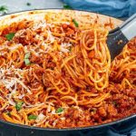 Spaghetti bolognese dengan saus daging melimpah dan taburan keju keju di dalam wajan besar yang sedang diangkat dengan penjepit makanan