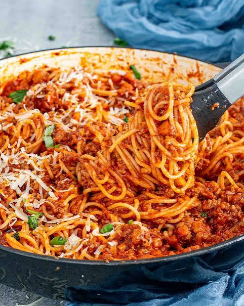 Spaghetti bolognese dengan saus daging melimpah dan taburan keju keju di dalam wajan besar yang sedang diangkat dengan penjepit makanan